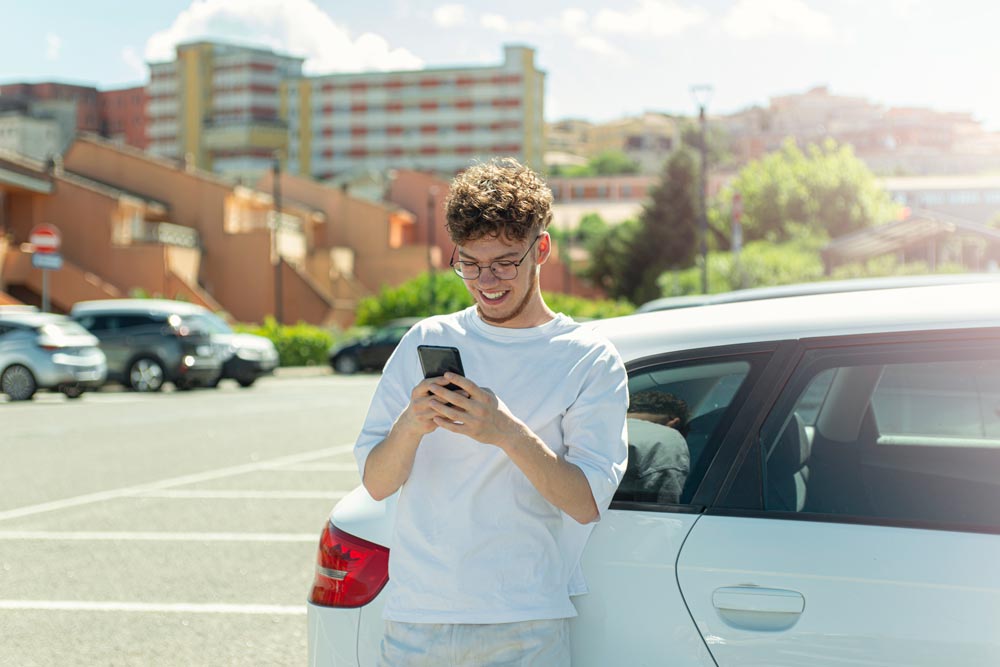 Tonåring står lutar mot bil med mobil i handen